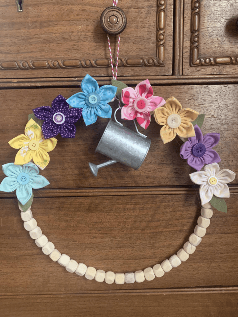 A DIY floral bead wreath featuring colorful fabric flowers and a mini watering can decoration