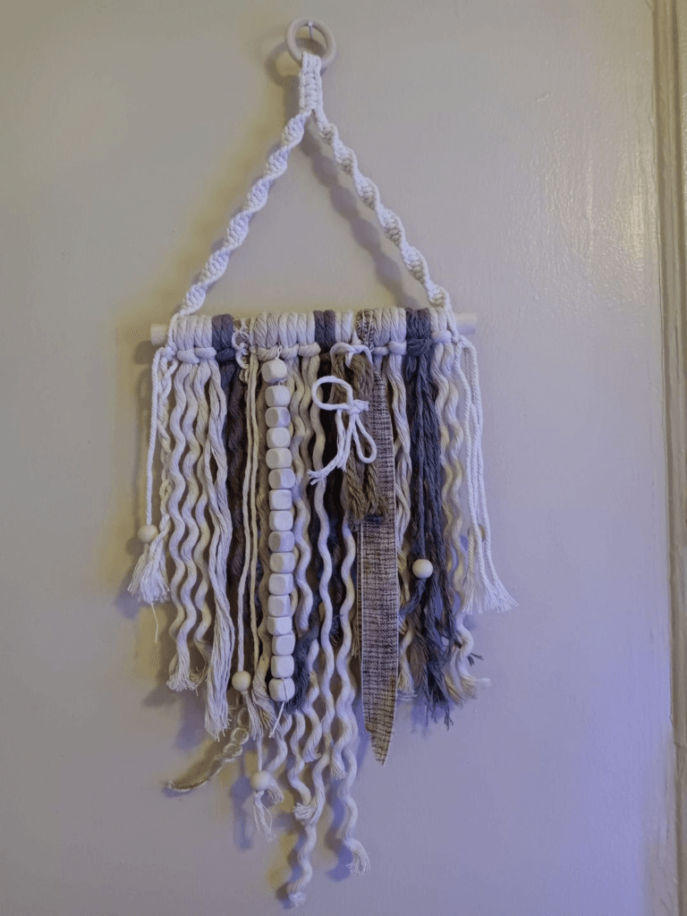 A DIY beaded wall hanging made with a towel holder, various ropes, ribbons, and wooden beads, creating a boho-inspired decor piece