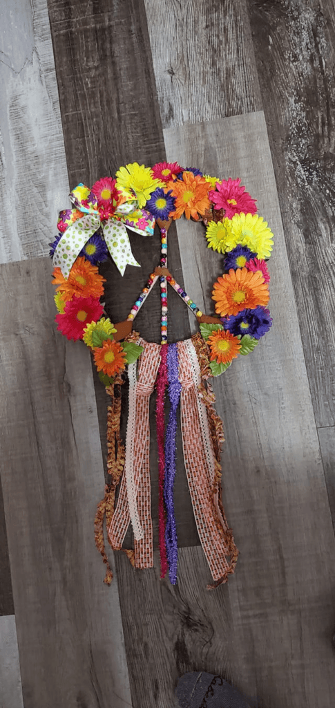 A colorful DIY wreath in the shape of a peace sign, decorated with bright faux flowers, beads, and cascading ribbons.