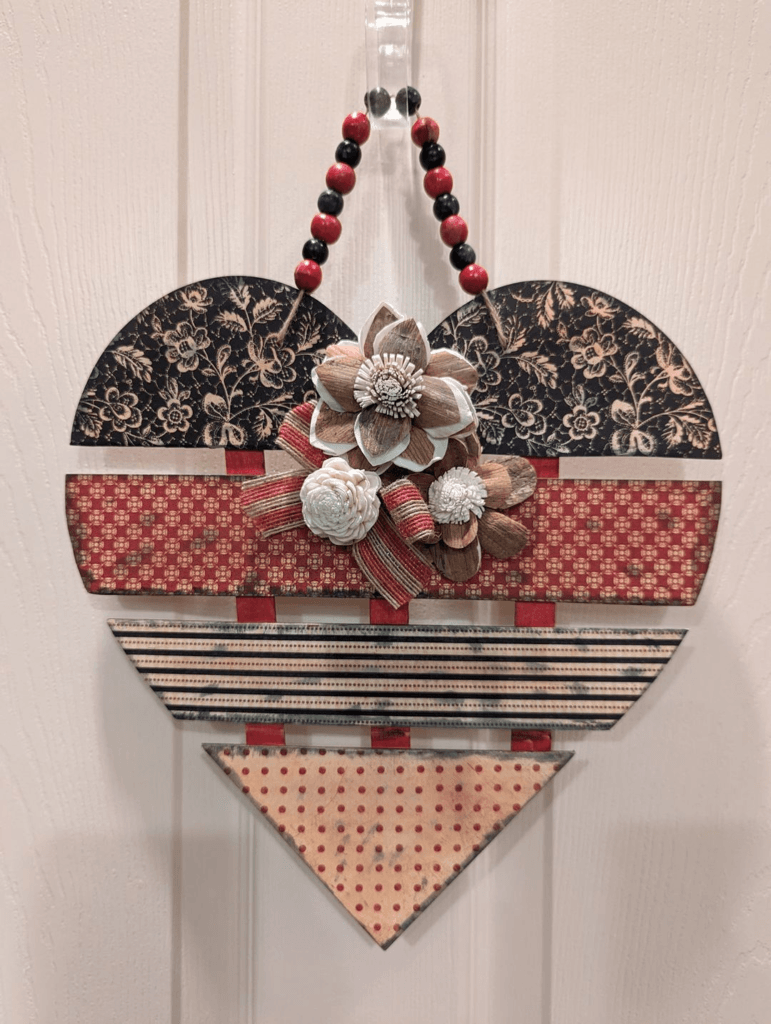 A handcrafted wooden heart door hanger decorated with vintage scrapbook paper, wooden flowers, and a beaded hanger, perfect for adding a rustic charm to your home decor.