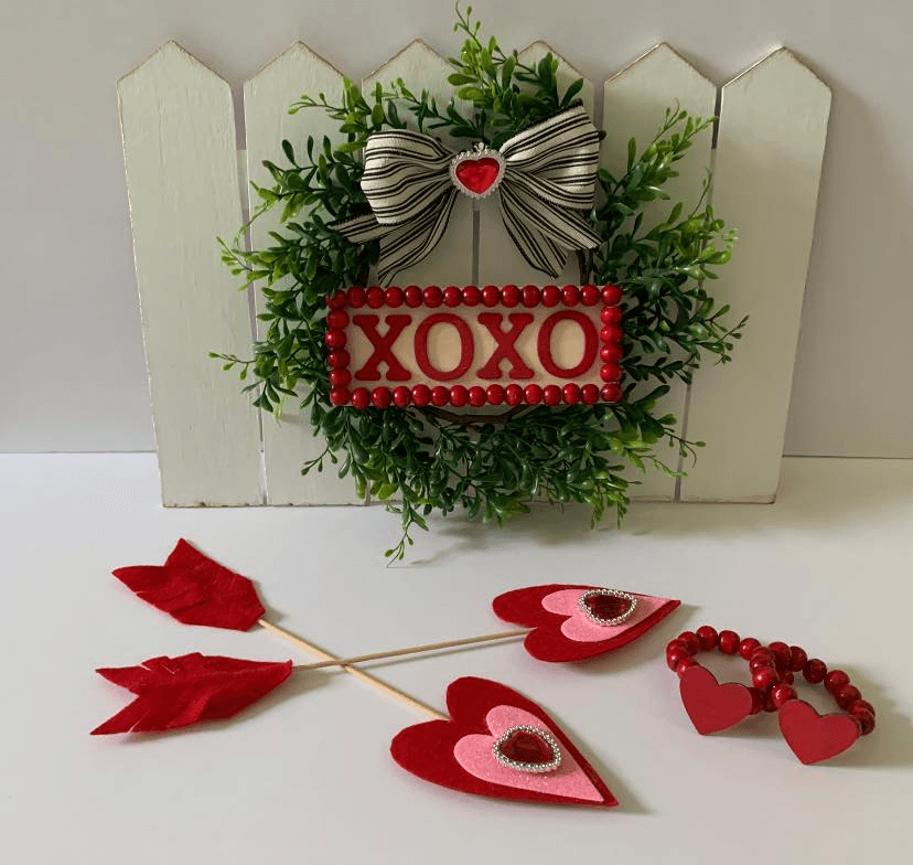 A mini Valentine’s wreath with "XOXO" sign on a white picket fence, accented with a bow.