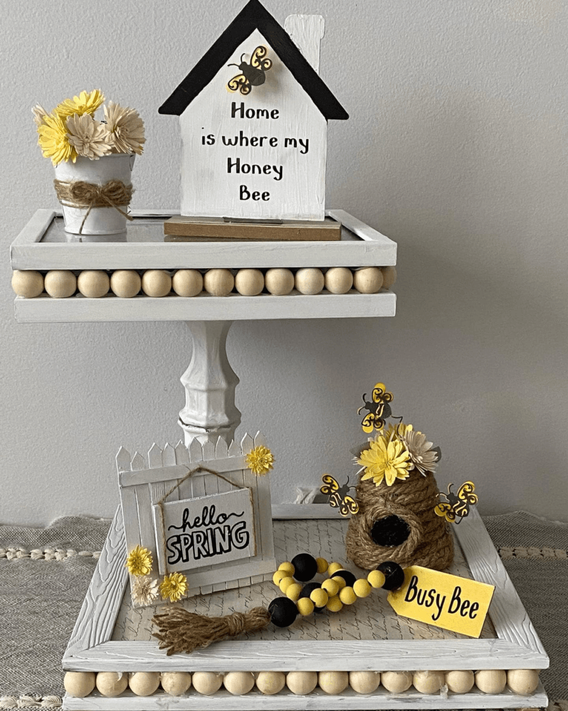 Spring-themed tiered tray decor with a bee house, 'Hello Spring' sign, and beehive, accented by wooden beads and yellow flowers.