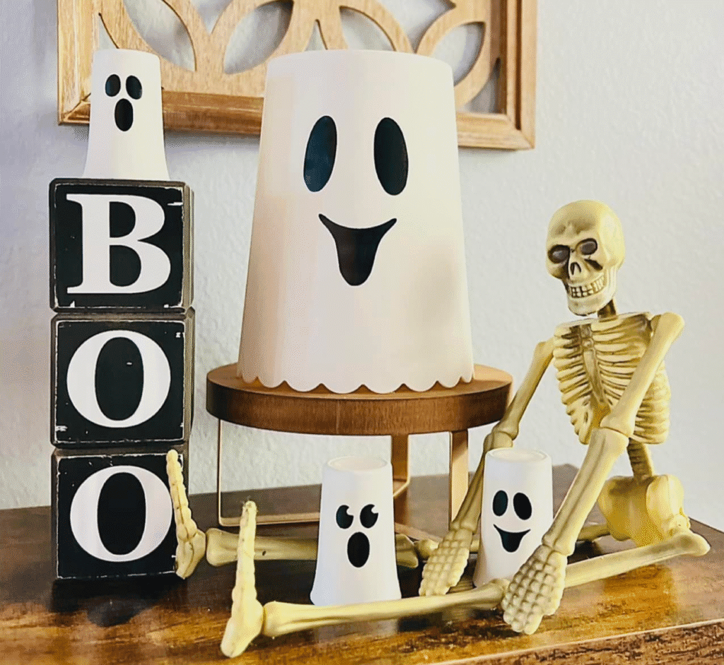 DIY Halloween ghost decor made from painted Dollar Tree canister and shot glasses with vinyl faces, paired with BOO blocks and a skeleton figure
