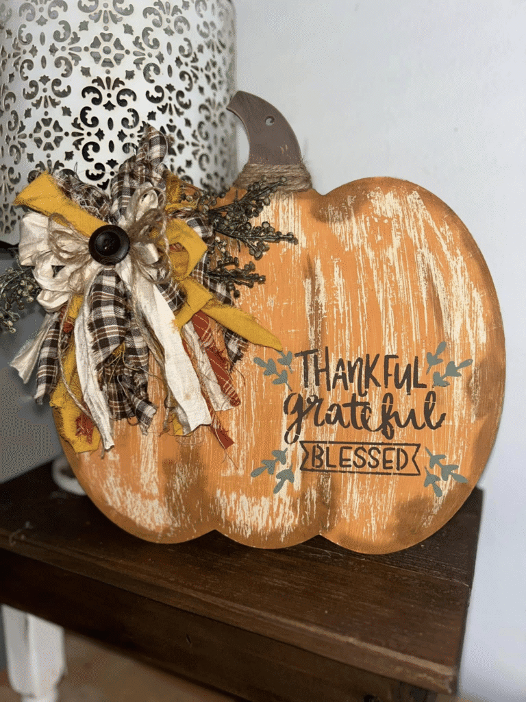DIY wooden pumpkin sign with distressed paint, plaid farmhouse bow, faux greenery, and the words Thankful Grateful Blessed
