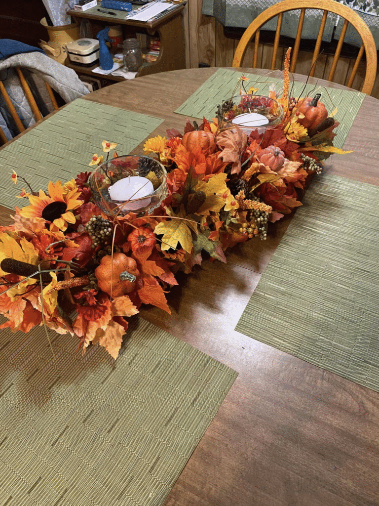 DIY fall centerpiece with faux pumpkins, autumn leaves, sunflowers, berries, and glass candle holders with tealights arranged on a dining table
