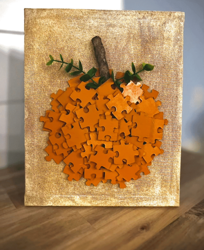 DIY pumpkin canvas made with orange-painted puzzle pieces, a stick stem, and greenery on a painted Dollar Tree canvas